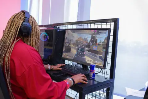 Graczka w czerwonej bluzie z warkoczami na głowie, słuchawkach JBL, gra w CS GO. Na biurku puszka Red Bulla.