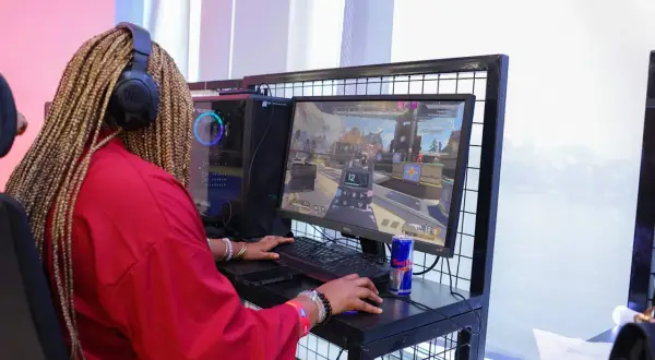 Graczka w czerwonej bluzie z warkoczami na głowie, słuchawkach JBL, gra w CS GO. Na biurku puszka Red Bulla.