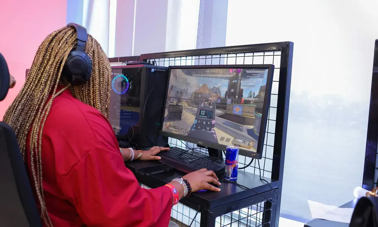 Graczka w czerwonej bluzie z warkoczami na głowie, słuchawkach JBL, gra w CS GO. Na biurku puszka Red Bulla.